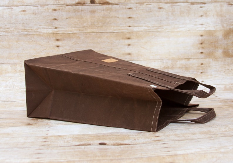 Pu&ograve; includere: Una borsa tote in tela marrone con una toppa in pelle e due manici. La borsa &egrave; piegata e appoggiata su una superficie di legno.