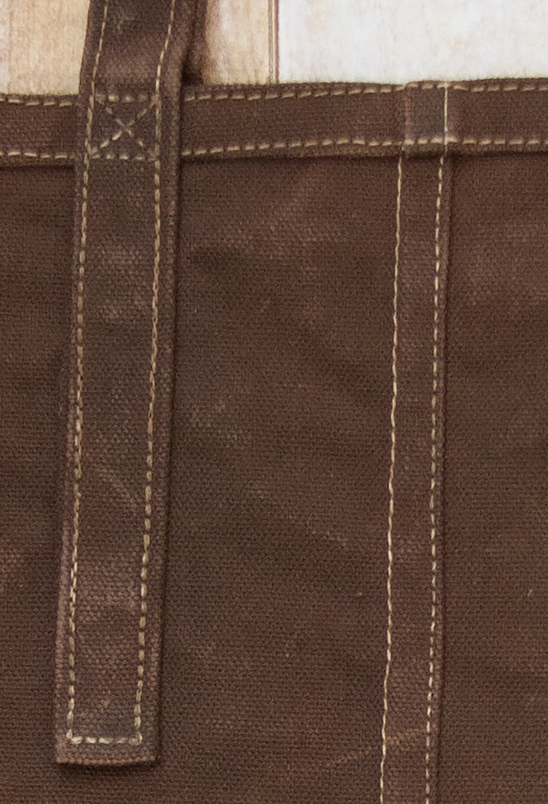 Pu&ograve; includere: Primo piano di una borsa tote in tela marrone con una tracolla in pelle marrone e cuciture bianche. La borsa ha un design semplice ed &egrave; realizzata in materiale resistente.