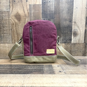 May include: A burgundy and olive green crossbody bag with a zippered front pocket and adjustable strap. The bag features a small rectangular patch with text. The bag is made of canvas material.