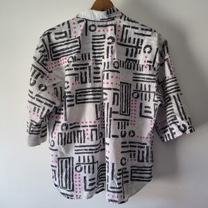 Puede incluir: Camisa blanca con un dise&ntilde;o geom&eacute;trico en negro y rosa. La camisa tiene mangas tres cuartos y un cuello cl&aacute;sico. El dise&ntilde;o presenta l&iacute;neas negras y puntos rosas dispuestos en un patr&oacute;n repetitivo.