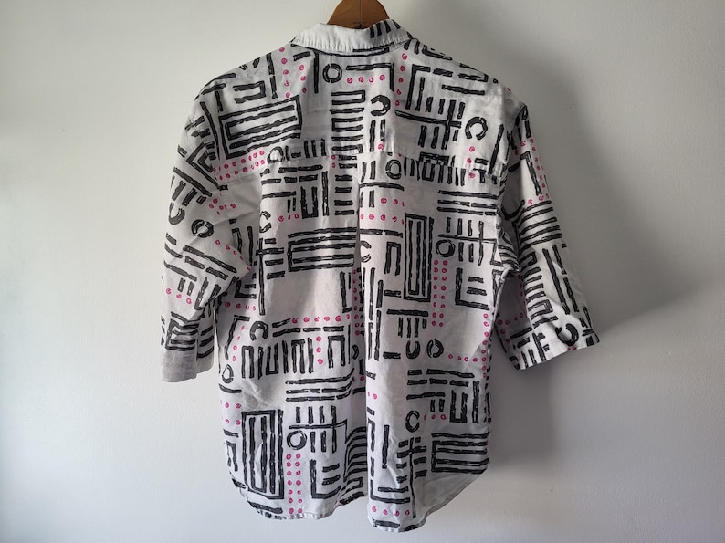 Puede incluir: Camisa blanca con un dise&ntilde;o geom&eacute;trico en negro y rosa. La camisa tiene mangas tres cuartos y un cuello cl&aacute;sico. El dise&ntilde;o presenta l&iacute;neas negras y puntos rosas dispuestos en un patr&oacute;n repetitivo.