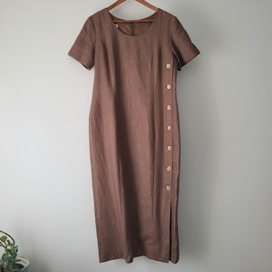 Puede incluir: Vestido de lino marrón con mangas cortas y cuello redondo. El vestido presenta una fila de botones en el lateral y una abertura en el dobladillo. El vestido está colgado de una percha de madera.
