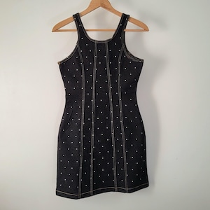 May include: A black denim dress with white polka dots and a scoop neckline hangs on a wooden hanger. The dress features a fitted silhouette, a small pocket, and contrasting tan stitching.