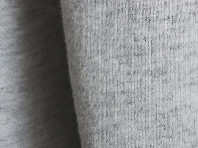 May include: A close-up view of light gray heathered fabric featuring a soft, fleecy texture. The material displays a subtle speckled pattern with varying shades of gray, creating a consistent, woven appearance across the entire surface of the textile.