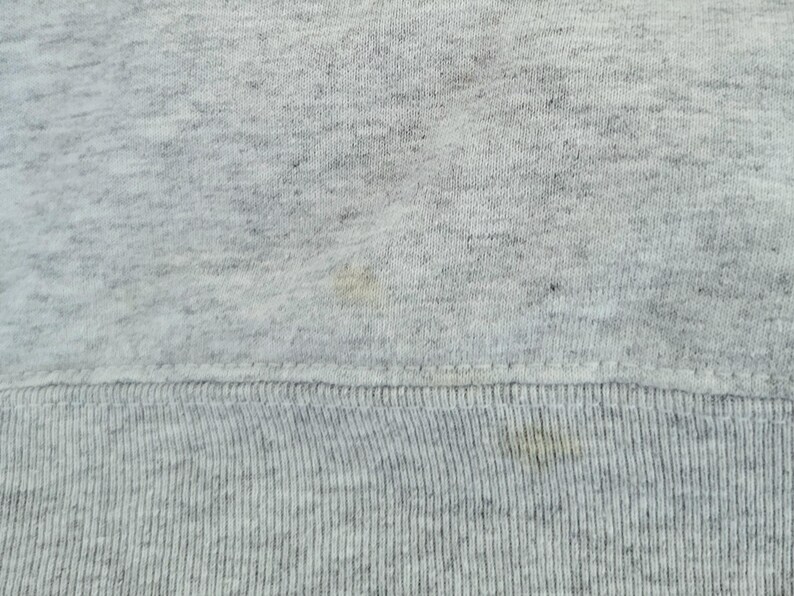 May include: A light gray heathered fabric with a textured knit surface. The material features a horizontal ribbed hemline at the bottom edge, with visible light yellow discoloration spots scattered across the fabric surface.