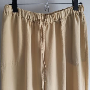 May include: A pair of tan trousers hanging on a black hanger. The trousers feature an elasticated waistband with a drawstring. The fabric looks to be a lightweight material, suitable for casual wear. The trousers are a solid colour with no visible patterns.