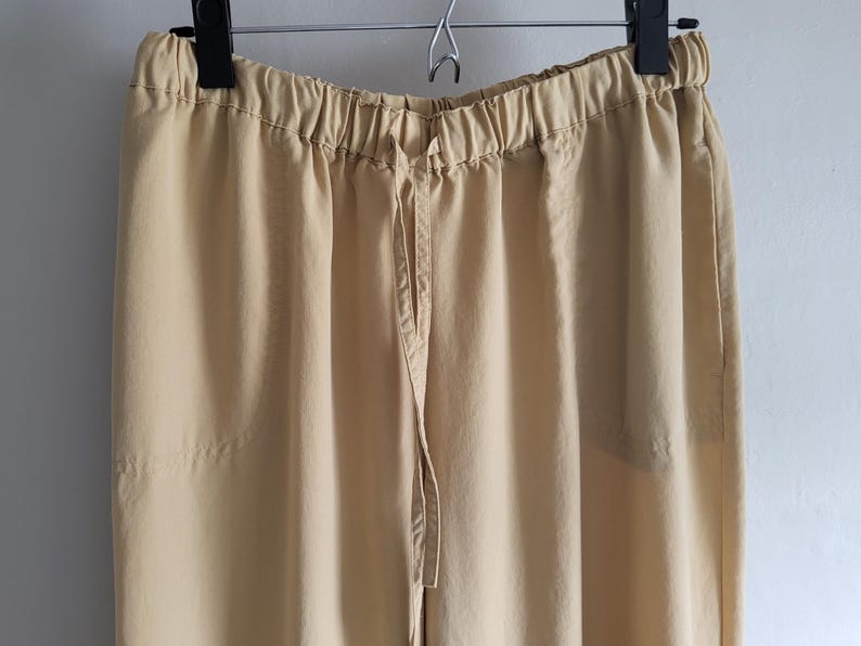 May include: A pair of tan trousers hanging on a black hanger. The trousers feature an elasticated waistband with a drawstring. The fabric looks to be a lightweight material, suitable for casual wear. The trousers are a solid colour with no visible patterns.