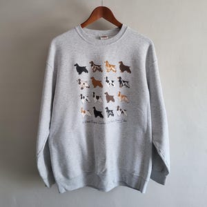 May include: A light gray crewneck sweatshirt featuring a grid of sixteen small illustrations of English Cocker Spaniel dogs in various coat colors. Below the dogs, the text reads A Good English Cocker Can Be A Best Color.