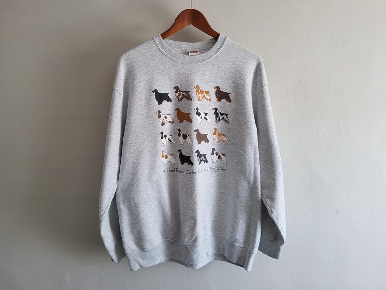 May include: A light gray crewneck sweatshirt featuring a grid of sixteen small illustrations of English Cocker Spaniel dogs in various coat colors. Below the dogs, the text reads A Good English Cocker Can Be A Best Color.