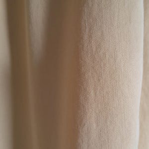 May include: Close-up of a section of light tan fabric. The material displays a subtle diagonal weave pattern, with soft folds and shadows adding depth. The texture appears smooth and slightly lustrous.