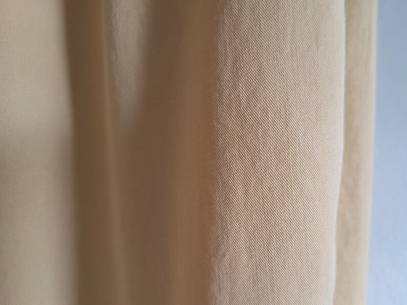 May include: Close-up of a section of light tan fabric. The material displays a subtle diagonal weave pattern, with soft folds and shadows adding depth. The texture appears smooth and slightly lustrous.