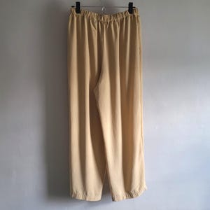May include: A pair of light tan, wide-leg trousers hanging on a metal hanger. The trousers have an elasticated waistband and a relaxed fit. The fabric appears to be a lightweight material, suitable for casual wear.