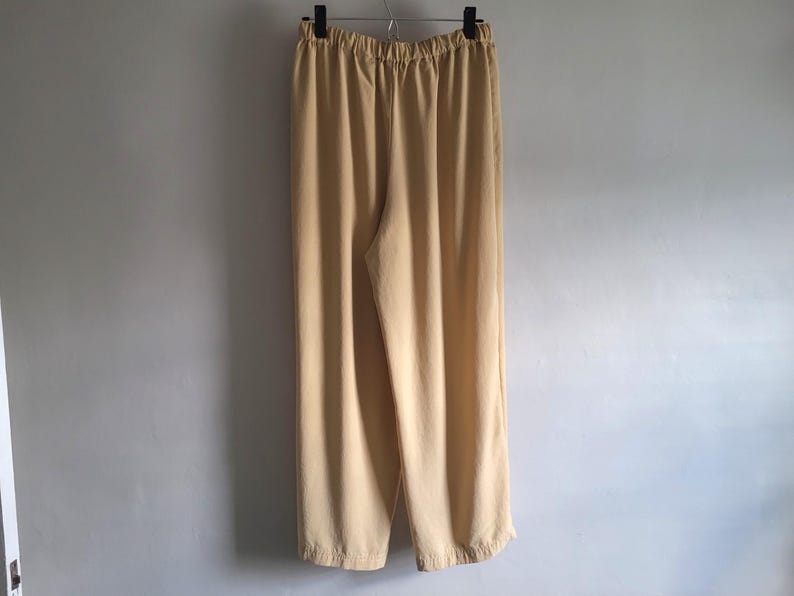 May include: A pair of light tan, wide-leg trousers hanging on a metal hanger. The trousers have an elasticated waistband and a relaxed fit. The fabric appears to be a lightweight material, suitable for casual wear.