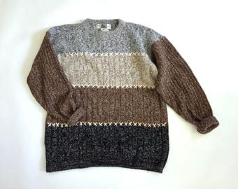 May include: A long-sleeve crew neck sweater featuring a color-blocked design with horizontal stripes in gray, beige, brown, and black. The sweater has a textured knit pattern and white cross-stitch detailing separating the color sections.