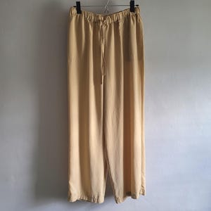 May include: Light tan wide-leg trousers hanging from a black hanger. The trousers feature a drawstring waist and a relaxed fit. The fabric looks lightweight, suitable for warmer weather. The trousers are against a plain, light-coloured wall. The length is approximately 100 cm.