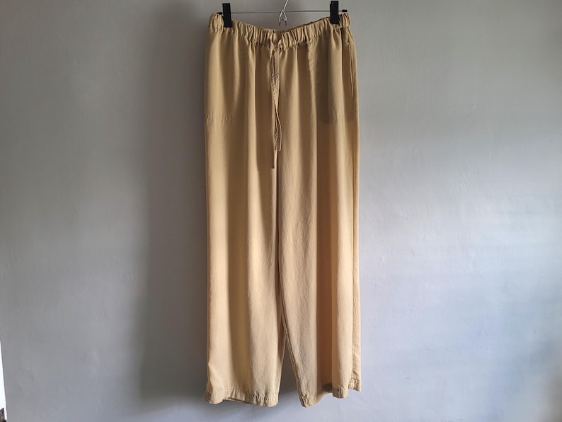 May include: Light tan wide-leg trousers hanging from a black hanger. The trousers feature a drawstring waist and a relaxed fit. The fabric looks lightweight, suitable for warmer weather. The trousers are against a plain, light-coloured wall. The length is approximately 100 cm.