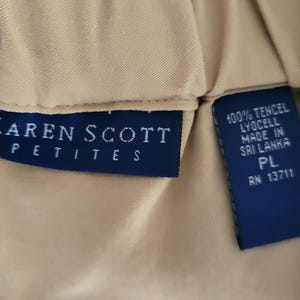 May include: Close-up of a tan garment with two navy blue tags. One tag reads "KAREN SCOTT PETITES" in white lettering. The other tag states "100% TENCEL LYOCELL MADE IN SRI LANKA PL RN 13711".