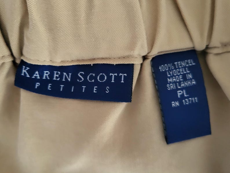 May include: Close-up of a tan garment with two navy blue tags. One tag reads "KAREN SCOTT PETITES" in white lettering. The other tag states "100% TENCEL LYOCELL MADE IN SRI LANKA PL RN 13711".