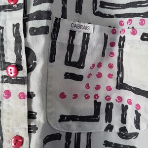 Puede incluir: Primer plano de una camisa blanca con un patr&oacute;n abstracto negro y detalles rosas. La camisa tiene un bolsillo con la marca "CABRAIS" y botones rosas.