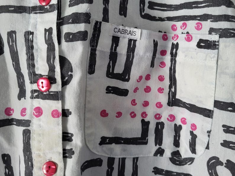 Puede incluir: Primer plano de una camisa blanca con un patr&oacute;n abstracto negro y detalles rosas. La camisa tiene un bolsillo con la marca "CABRAIS" y botones rosas.