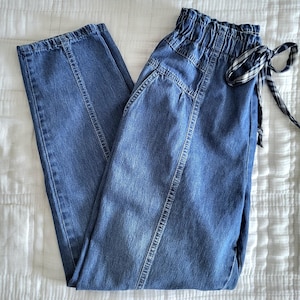May include: Blue denim pants with a high, elasticized waist and a drawstring with a black and white plaid pattern. The pants have a straight leg design and vertical seam details. The pants are folded on a white surface.