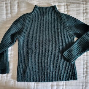 Puede incluir: Un suéter de punto acanalado verde azulado con cuello alto y mangas largas. El suéter está hecho de un material suave y texturizado y se muestra sobre una superficie blanca y texturizada.