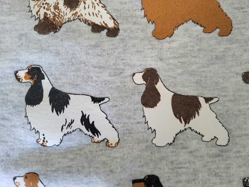 May include: A light gray fabric featuring a repeating pattern of spaniel dogs in various color combinations, including solid brown, white with black patches, and white with brown patches, all depicted in a side profile standing position.
