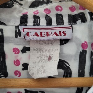 Puede incluir: Primer plano de una camisa blanca con un estampado abstracto en negro y rosa. La camisa tiene una etiqueta que dice "CABRAIS" e incluye instrucciones de lavado. La camisa est&aacute; colgada en una percha de madera.