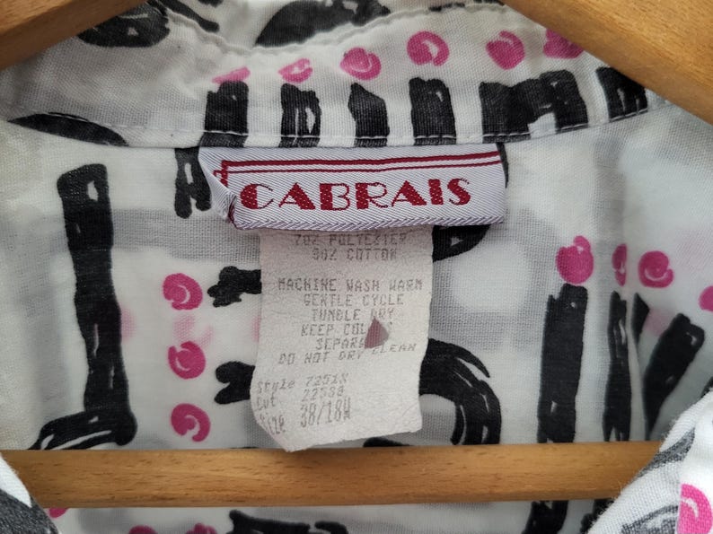 Puede incluir: Primer plano de una camisa blanca con un estampado abstracto en negro y rosa. La camisa tiene una etiqueta que dice "CABRAIS" e incluye instrucciones de lavado. La camisa est&aacute; colgada en una percha de madera.