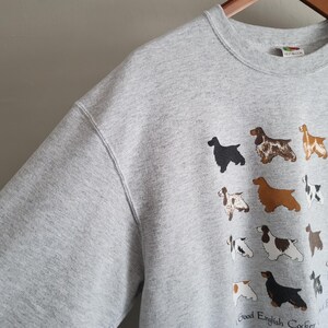 May include: A light gray crewneck sweatshirt featuring a graphic print of various English Cocker Spaniel dogs in different coat colors and patterns. The text at the bottom reads Good English Cocker and Bad Color, with a small brand tag at the neckline.
