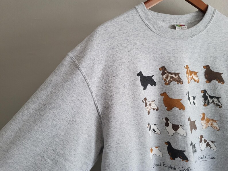 May include: A light gray crewneck sweatshirt featuring a graphic print of various English Cocker Spaniel dogs in different coat colors and patterns. The text at the bottom reads Good English Cocker and Bad Color, with a small brand tag at the neckline.