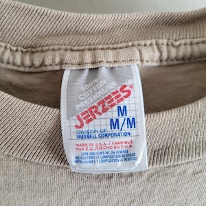 K&ouml;nnte beinhalten: Nahaufnahme eines beigefarbenen T-Shirt-Kragens mit einem wei&szlig;en Etikett. Das Etikett zeigt das "JERZEES"-Logo in Rot und Wei&szlig; sowie die Gr&ouml;&szlig;enangaben "M" und "M/M". Das Etikett gibt auch an, dass das T-Shirt aus 100 % Baumwolle besteht und in den USA hergestellt wurde.
