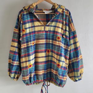Op de afbeelding: Een geruit shirt met capuchon in geel, blauw en rood met geborduurde zonnebloemen. Het shirt heeft een trekkoord in de taille en lange mouwen. Het hangt aan een houten hanger.