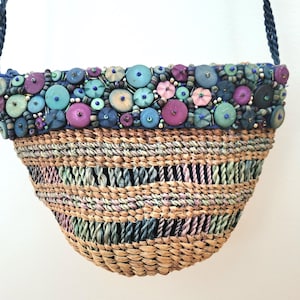 Può includere: Borsa a tracolla intrecciata con cinturino blu navy intrecciato. La parte superiore è decorata con perline e bottoni blu, viola e verdi. Il corpo della borsa è realizzato con fibre naturali intrecciate. Un accessorio unico.