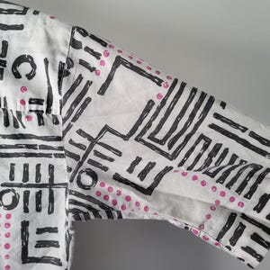 Puede incluir: Camisa blanca de manga corta con un patr&oacute;n geom&eacute;trico negro y peque&ntilde;os puntos rosas. El dise&ntilde;o presenta una mezcla de l&iacute;neas, rect&aacute;ngulos y c&iacute;rculos, creando un aspecto audaz y abstracto. Se ve la manga de la camisa.