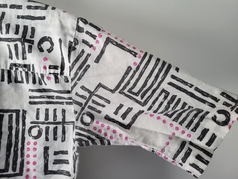 Puede incluir: Camisa blanca de manga corta con un patr&oacute;n geom&eacute;trico negro y peque&ntilde;os puntos rosas. El dise&ntilde;o presenta una mezcla de l&iacute;neas, rect&aacute;ngulos y c&iacute;rculos, creando un aspecto audaz y abstracto. Se ve la manga de la camisa.