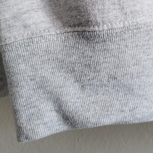 May include: A light gray heathered sweatshirt featuring a ribbed knit cuff at the sleeve end. The fabric displays a speckled texture throughout, with a horizontal seam connecting the ribbed cuff to the main body of the garment.