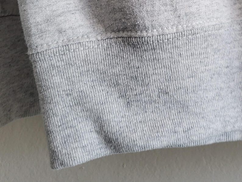 May include: A light gray heathered sweatshirt featuring a ribbed knit cuff at the sleeve end. The fabric displays a speckled texture throughout, with a horizontal seam connecting the ribbed cuff to the main body of the garment.
