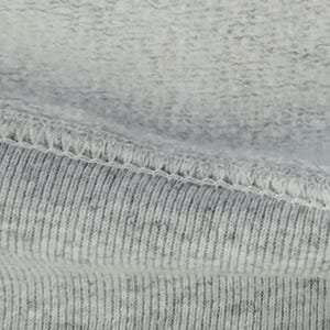 May include: A light gray heathered fabric featuring a soft, fleecy interior texture contrasted against a ribbed exterior surface. The two distinct fabric textures are joined together by a visible, reinforced white overlock stitch seam.