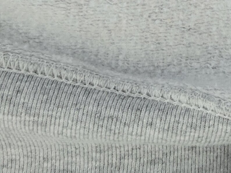 May include: A light gray heathered fabric featuring a soft, fleecy interior texture contrasted against a ribbed exterior surface. The two distinct fabric textures are joined together by a visible, reinforced white overlock stitch seam.