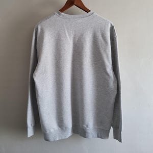 May include: A light gray crewneck sweatshirt featuring long sleeves and ribbed cuffs. The garment is displayed from the back, showing a solid, heathered fabric texture throughout the entire piece, hanging on a wooden hanger against a plain wall.