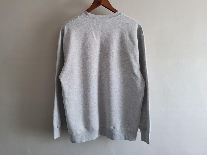 May include: A light gray crewneck sweatshirt featuring long sleeves and ribbed cuffs. The garment is displayed from the back, showing a solid, heathered fabric texture throughout the entire piece, hanging on a wooden hanger against a plain wall.
