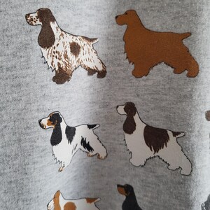 May include: A light gray fabric featuring a repeating pattern of various spaniel dog breeds. The illustrations include dogs with brown and white spotted coats, solid brown coats, and black and white markings, all depicted in a standing profile view.