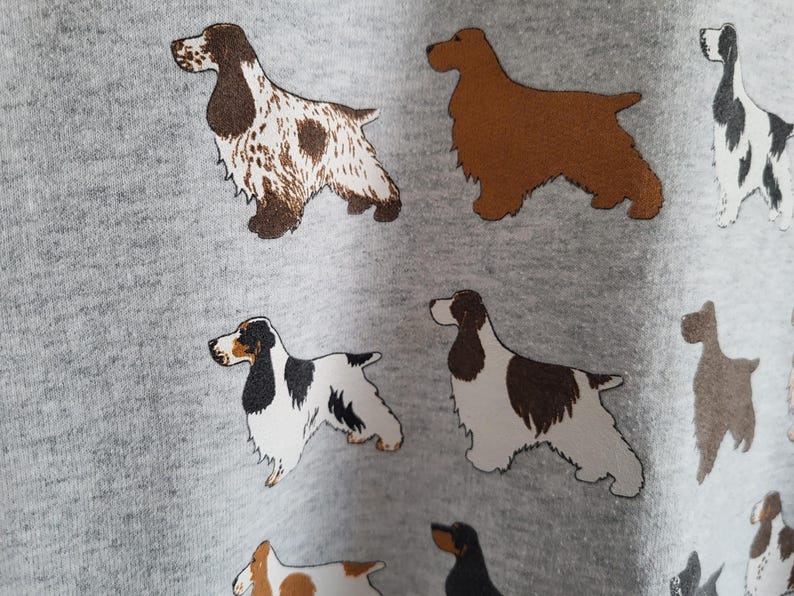 May include: A light gray fabric featuring a repeating pattern of various spaniel dog breeds. The illustrations include dogs with brown and white spotted coats, solid brown coats, and black and white markings, all depicted in a standing profile view.