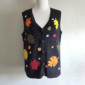May include: A black knit vest featuring a V-neckline and a front button closure. The garment is decorated with embroidered and appliqued autumn leaves in shades of orange, yellow, red, and olive green, accented with small white floral details.