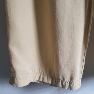 May include: Close-up of beige trousers. The fabric looks lightweight, with seams and a hem at the bottom. The trousers are slightly creased, suggesting a soft fabric. The colour is a light, neutral tone.