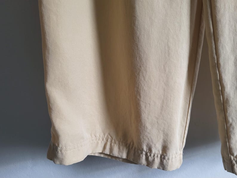 May include: Close-up of beige trousers. The fabric looks lightweight, with seams and a hem at the bottom. The trousers are slightly creased, suggesting a soft fabric. The colour is a light, neutral tone.