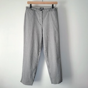 Può includere: Un paio di pantaloni a quadri bianchi e neri appesi a una gruccia di legno. I pantaloni hanno un taglio dritto e una chiusura con bottone e cerniera. Il tessuto presenta un piccolo motivo ripetuto.