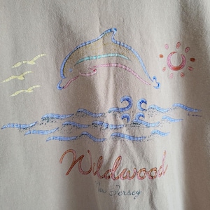 K&ouml;nnte beinhalten: Beiges Sweatshirt mit einem gestickten Design eines Delfins, der &uuml;ber Wellen springt, mit Sonne und M&ouml;wen. Das Wort "Wildwood" steht in roter Schrift, darunter "New Jersey". Der Delfin hat bunte Streifen.