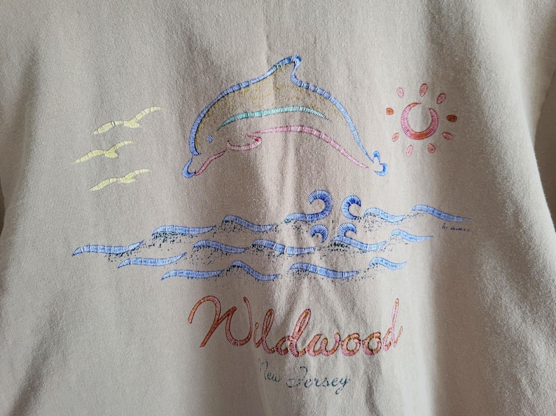 K&ouml;nnte beinhalten: Beiges Sweatshirt mit einem gestickten Design eines Delfins, der &uuml;ber Wellen springt, mit Sonne und M&ouml;wen. Das Wort "Wildwood" steht in roter Schrift, darunter "New Jersey". Der Delfin hat bunte Streifen.